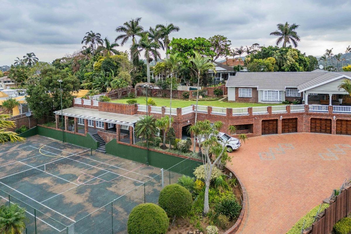 Aerial view of a large residential property featuring a private tennis court, extensive brick driveway, triple garage, and a raised terrace area.