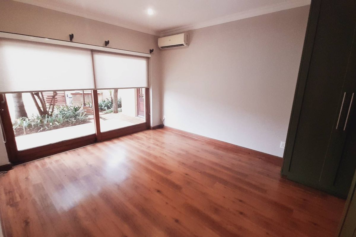 A bright bedroom featuring wood-look laminate flooring, a built-in wardrobe, and a wall-mounted air conditioning unit with sliding door access to the garden.