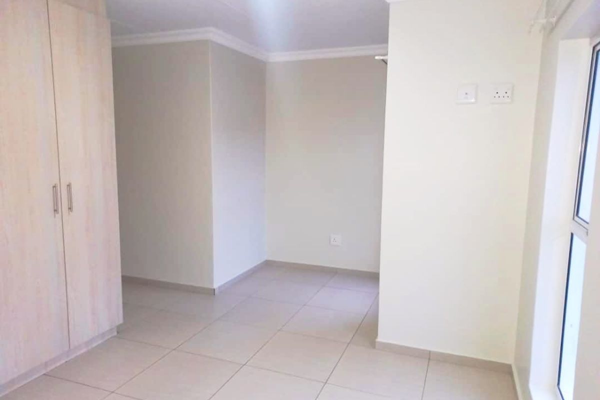 A bright, empty bedroom featuring neutral tiled flooring, built-in wooden cupboards, and large windows allowing for ample natural light.