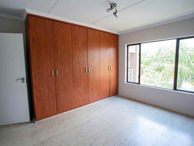 A bright, empty bedroom featuring extensive built-in wooden cupboards, light-toned laminate flooring, and large windows providing natural light.