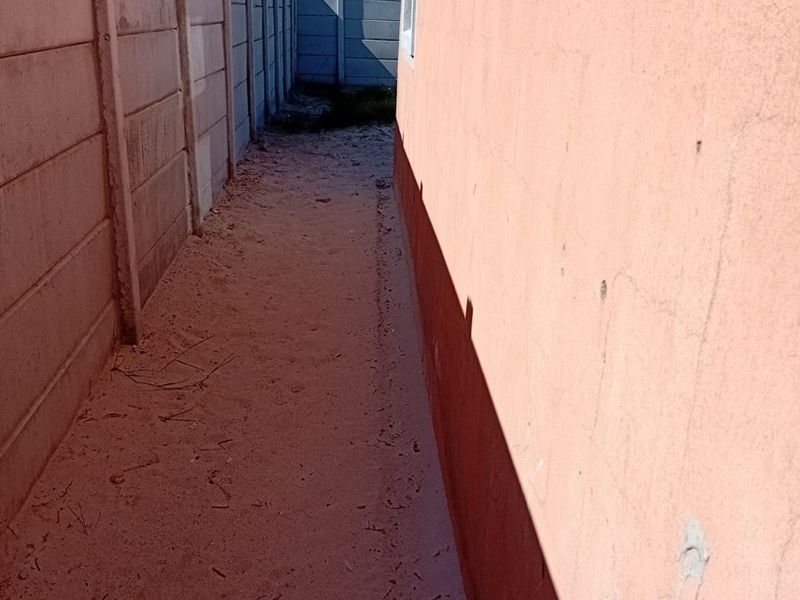 A narrow side passage between the house exterior wall and a precast concrete boundary wall, featuring an unpaved ground surface.