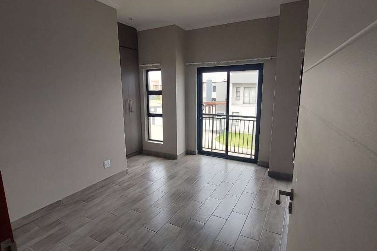 A bright, empty bedroom featuring modern wood-look tiled flooring, built-in cupboards, and a sliding glass door leading to a balcony.