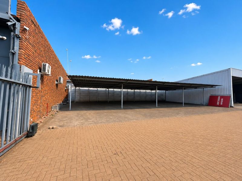 A spacious outdoor parking area featuring a large covered carport, paved driveway, and secure perimeter walling, suitable for commercial or residential vehicle storage.