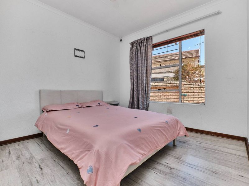A functional bedroom featuring light-toned laminate flooring, a window with security bars, and simple white walls.
