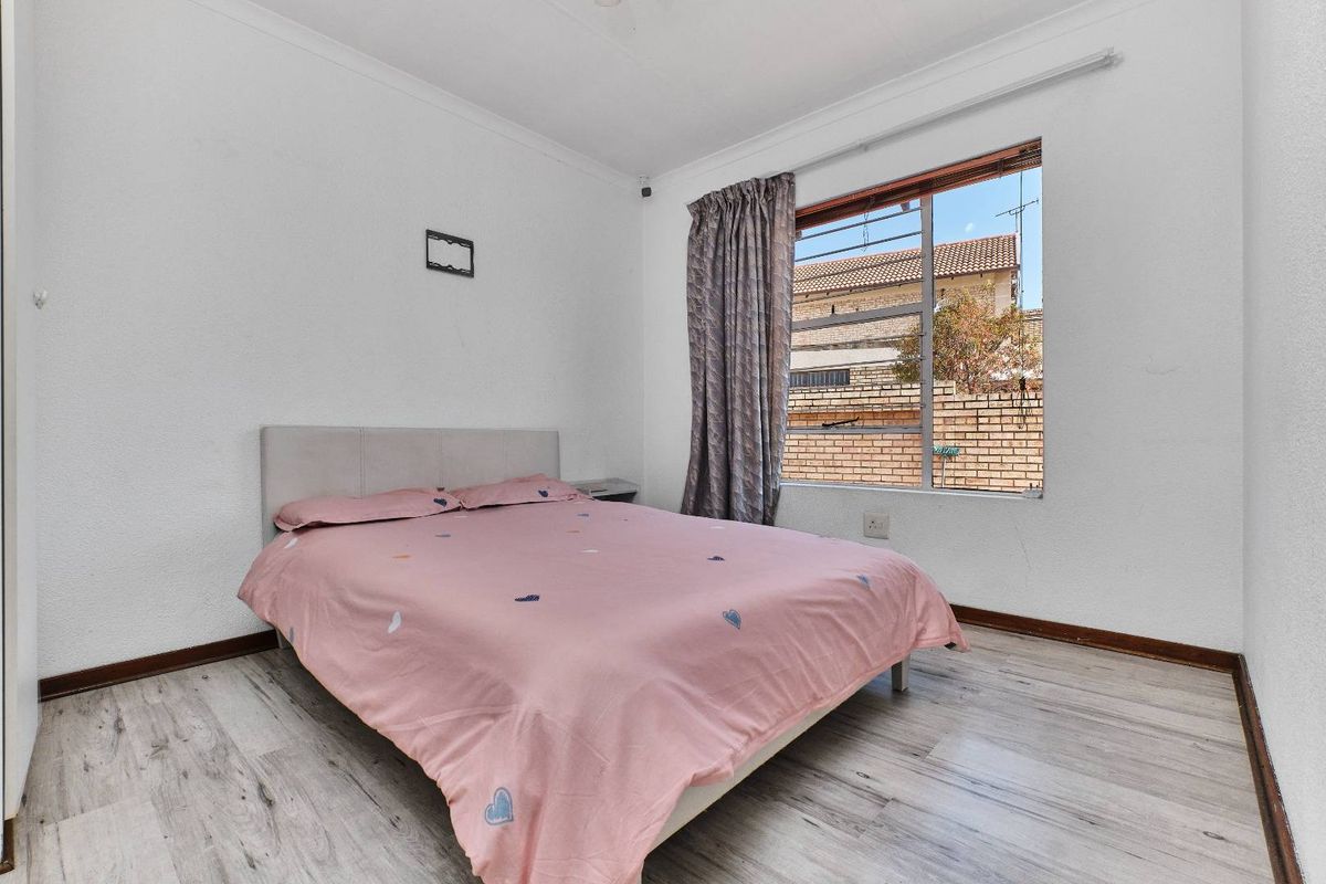A functional bedroom featuring light-toned laminate flooring, a window with security bars, and simple white walls.