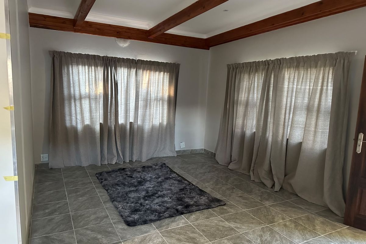A bright, empty living room featuring exposed timber ceiling beams, neutral tiled flooring, and large windows dressed with floor-to-ceiling curtains.