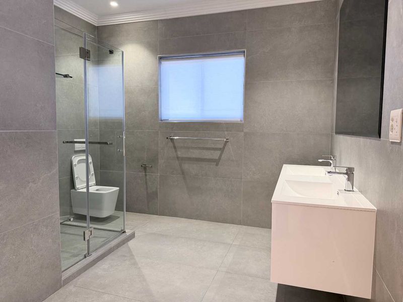 Modern bathroom featuring a double vanity, wall-mounted toilet, and a spacious glass-enclosed shower with contemporary grey tiling throughout.