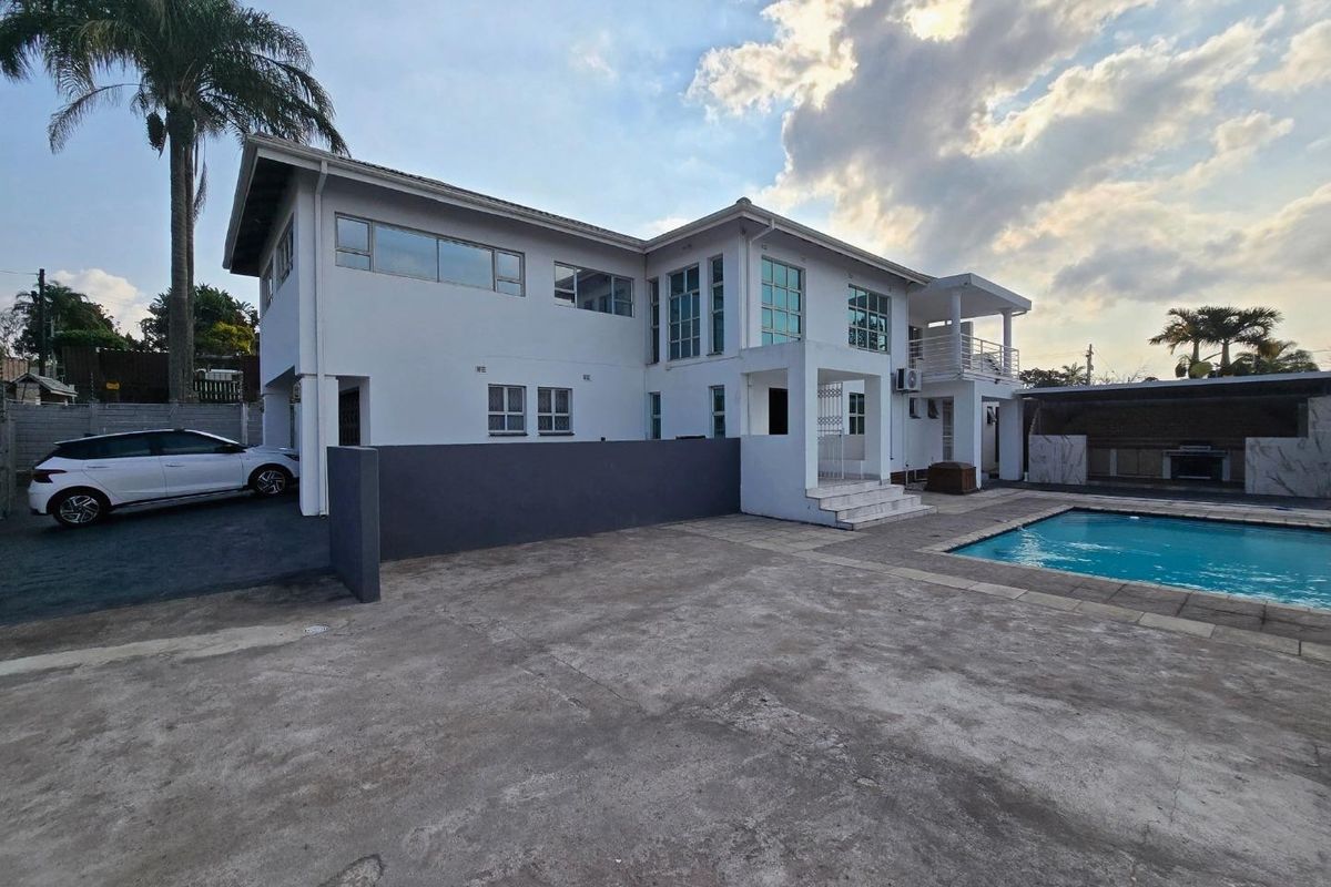 Rear exterior view of a two-story house featuring a swimming pool, covered patio area with built-in braai, and a balcony.