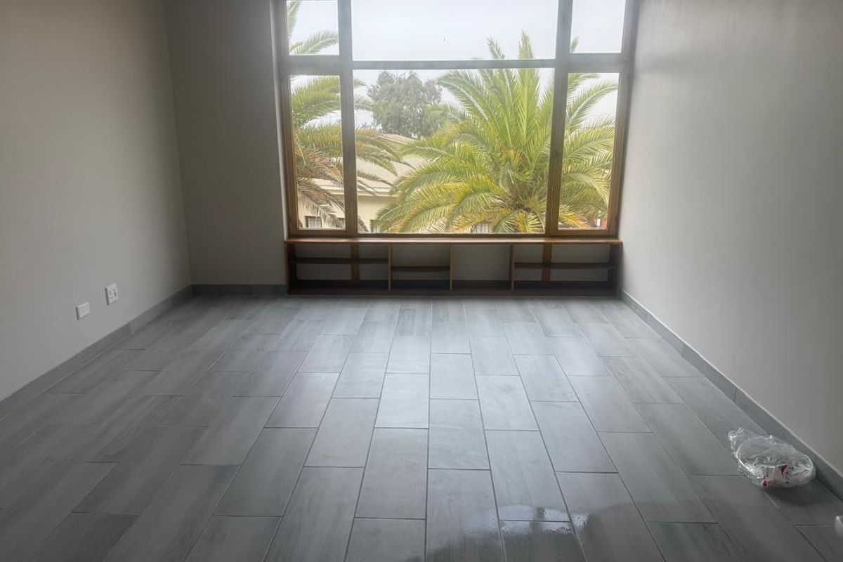 A bright, empty bedroom featuring modern grey tiled flooring, a large window with garden views, and integrated low-level built-in shelving.