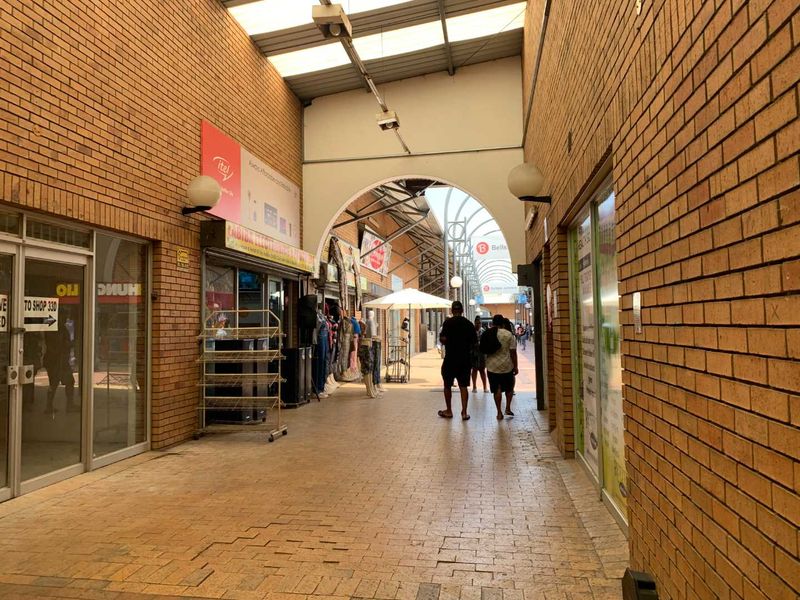 A covered pedestrian walkway within a retail center, featuring brick walls, paved flooring, and multiple glass-fronted shop units.