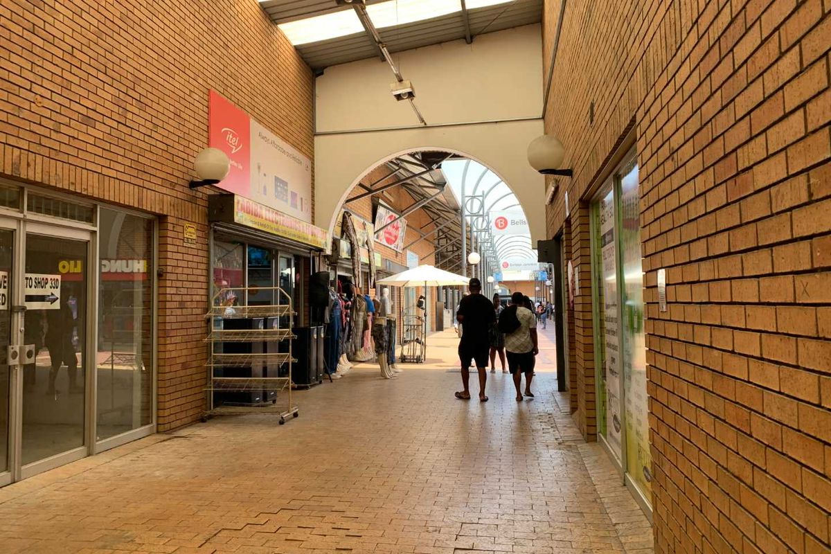 A covered pedestrian walkway within a retail center, featuring brick walls, paved flooring, and multiple glass-fronted shop units.