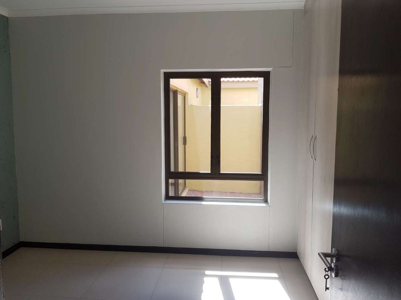 A clean, empty bedroom featuring neutral tiled flooring, built-in cupboards, and an aluminium window frame overlooking a small courtyard.