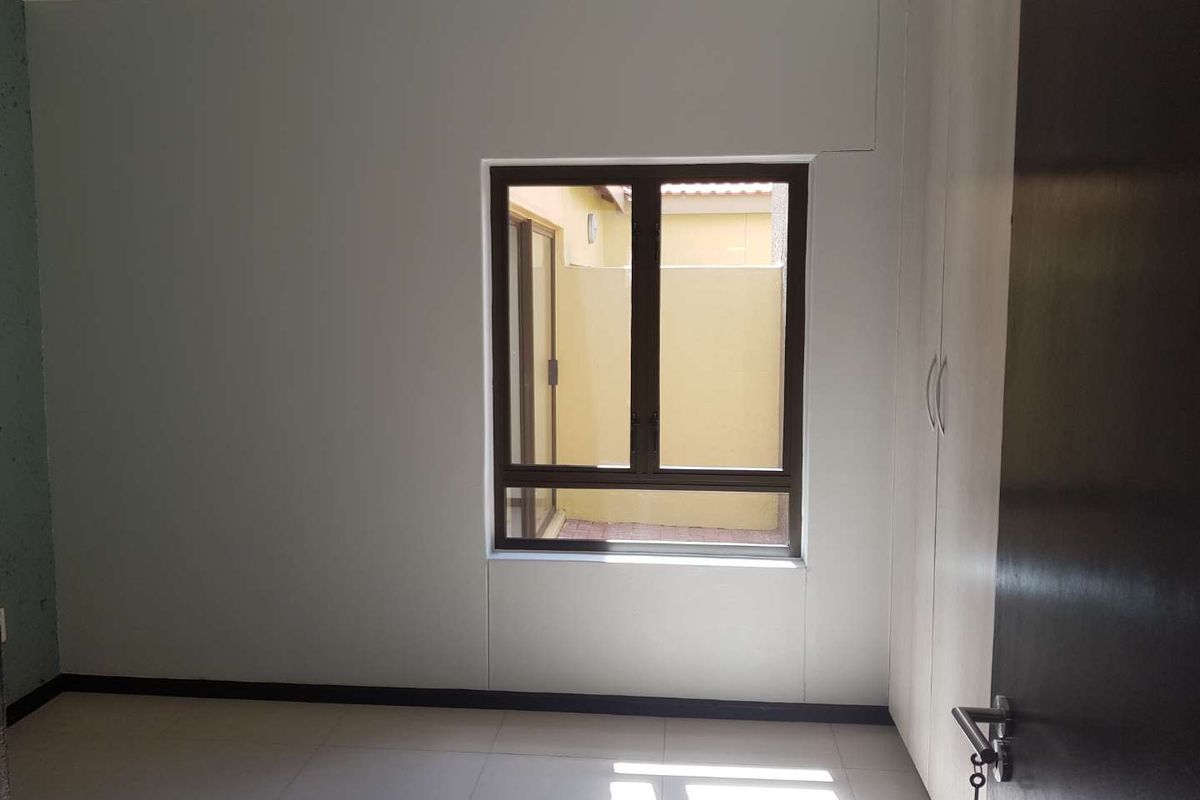 A clean, empty bedroom featuring neutral tiled flooring, built-in cupboards, and an aluminium window frame overlooking a small courtyard.