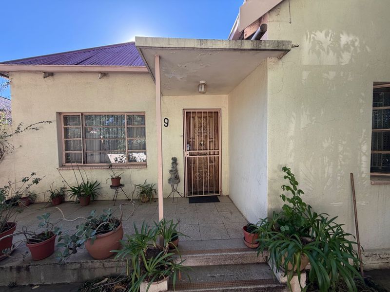 Front entrance of a house featuring a tiled porch, metal security gate, and steel frame windows, showing signs of exterior wear.