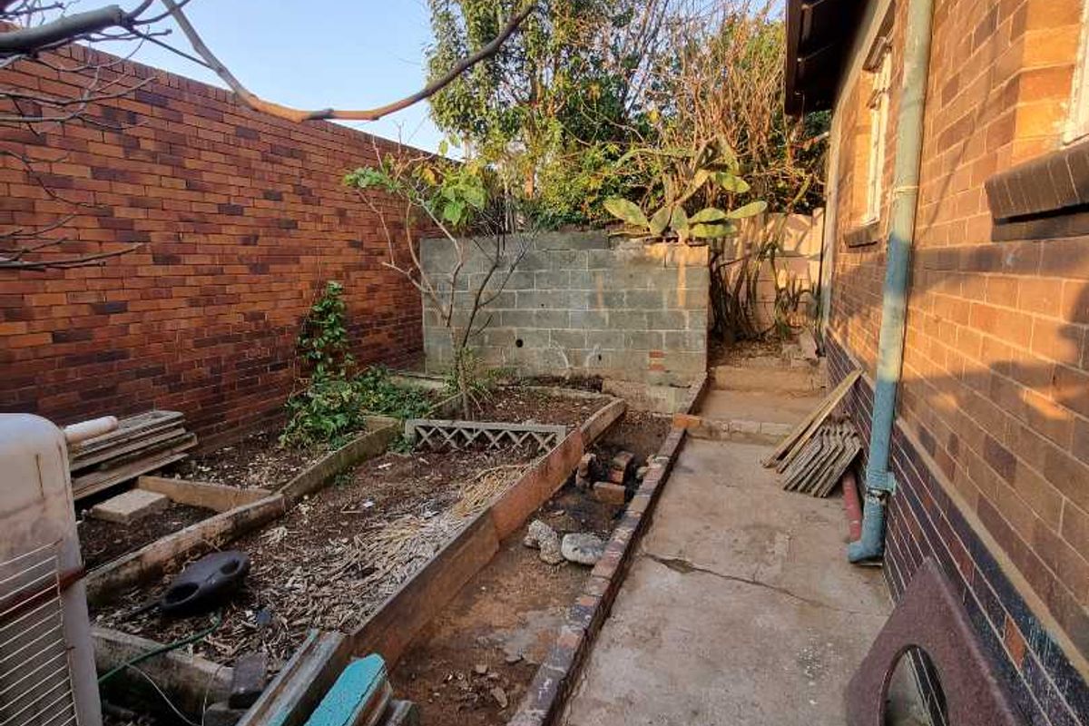 A rear exterior area featuring a concrete pathway, brick boundary walls, and raised garden beds in need of maintenance and landscaping.
