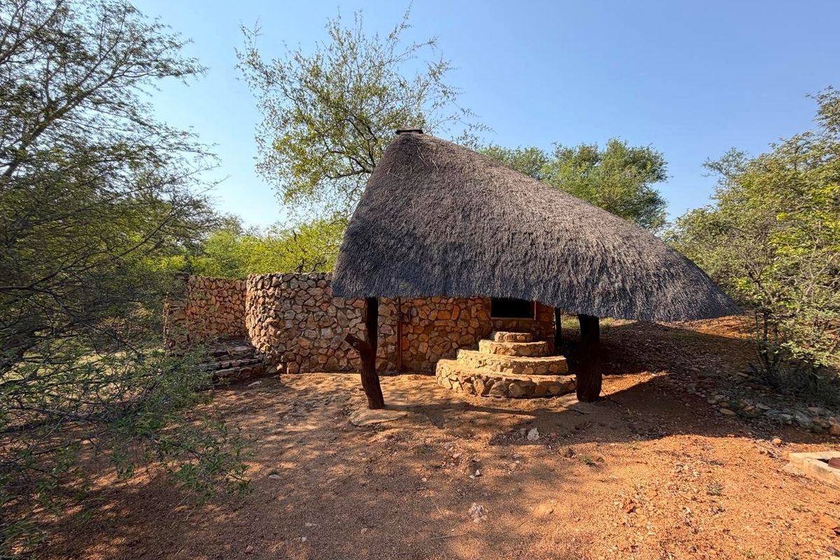 A rustic outdoor structure featuring a traditional thatched roof, stone masonry walls, and stone steps, set within a natural bushveld environment.