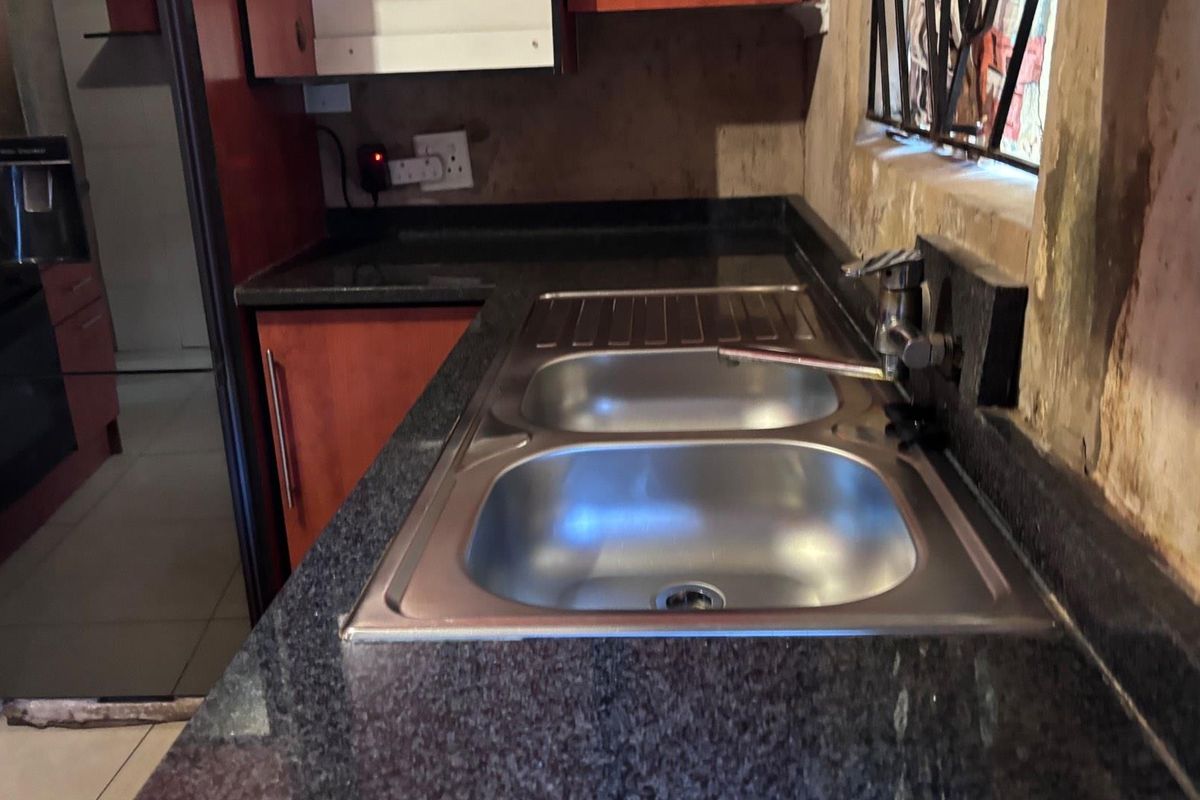 A kitchen area featuring dark granite countertops, a double stainless steel sink, and wooden cabinetry, showing signs of wear and wall surface damage.