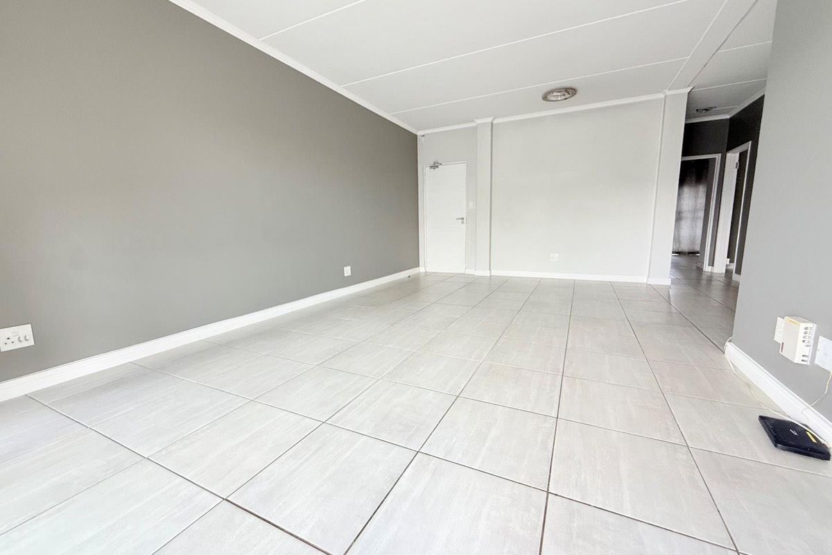 A spacious, empty living area featuring neutral grey and white walls, tiled flooring, and recessed ceiling lighting, offering a clean and versatile living space.
