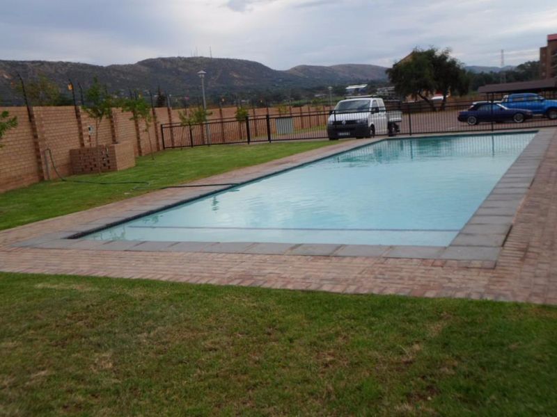 A well-maintained communal swimming pool area featuring brick paving, a lawn, and secure perimeter fencing with mountain views.