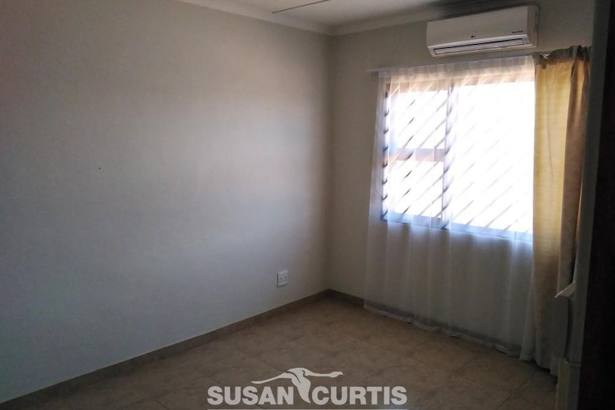 A simple, empty bedroom featuring tiled flooring, a wall-mounted air conditioning unit, and a window with security bars.