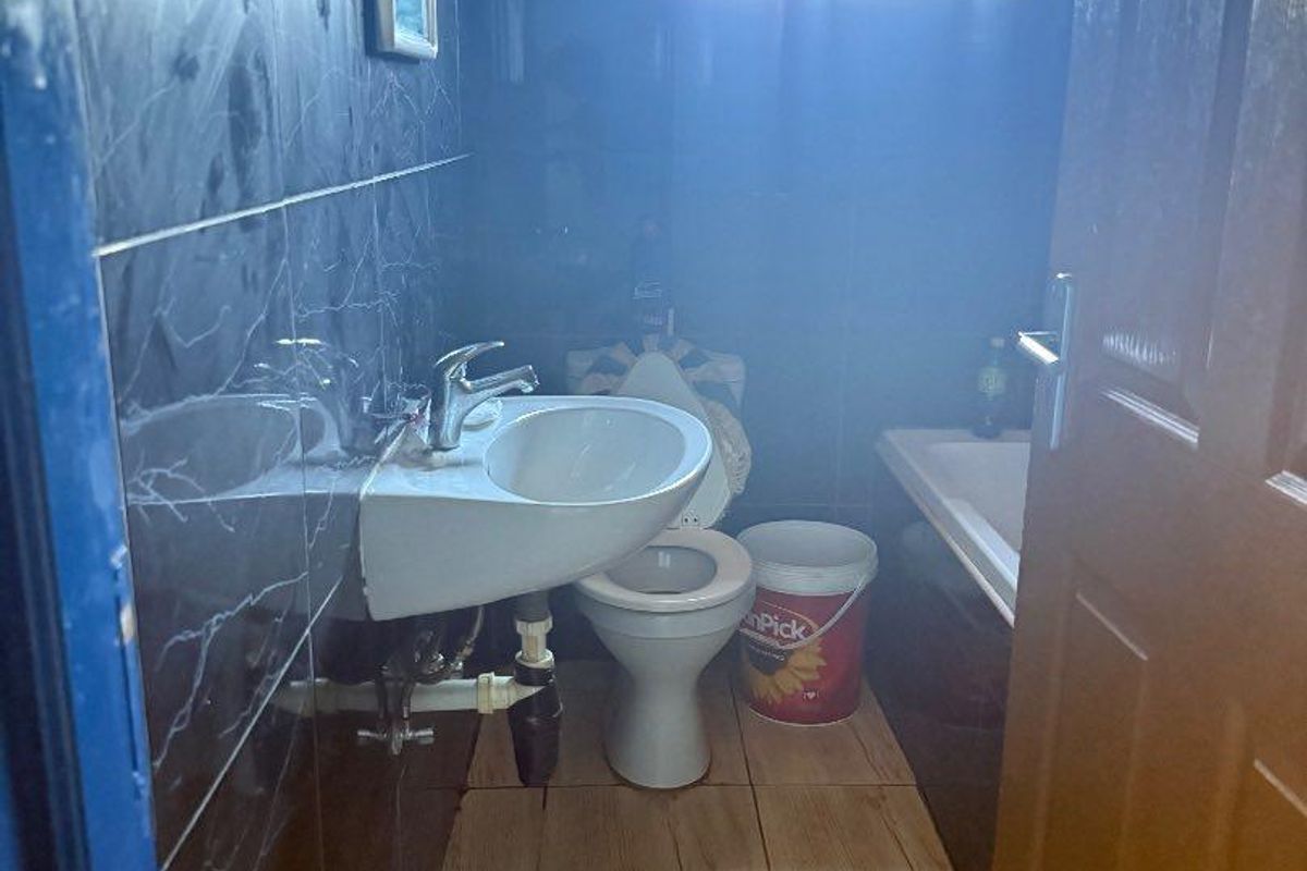A compact bathroom featuring dark tiled walls, a wall-mounted basin with exposed plumbing, a toilet, and a bathtub with a window.