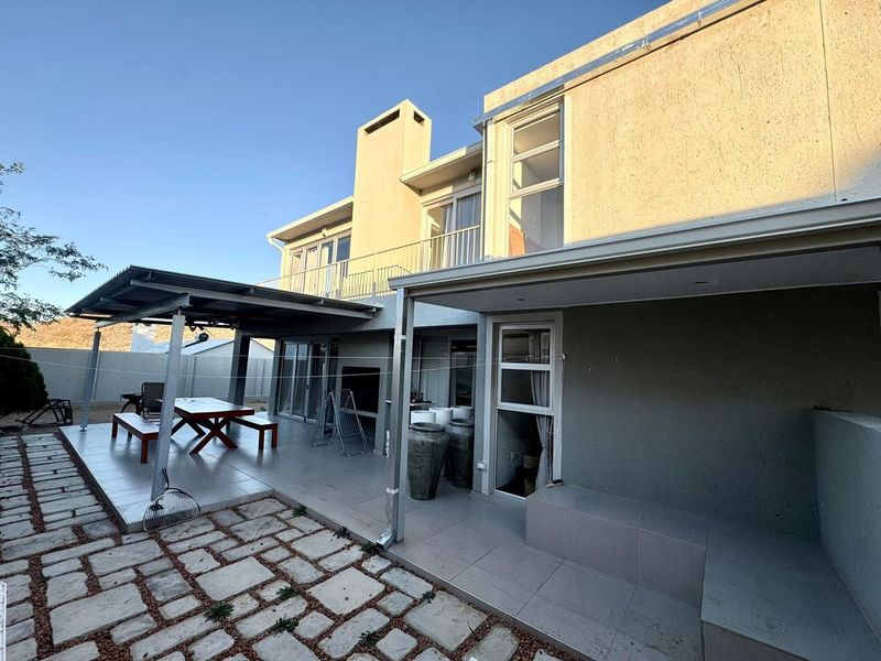 Rear exterior view featuring a spacious covered patio with built-in braai, tiled flooring, and access to an upper-level balcony.
