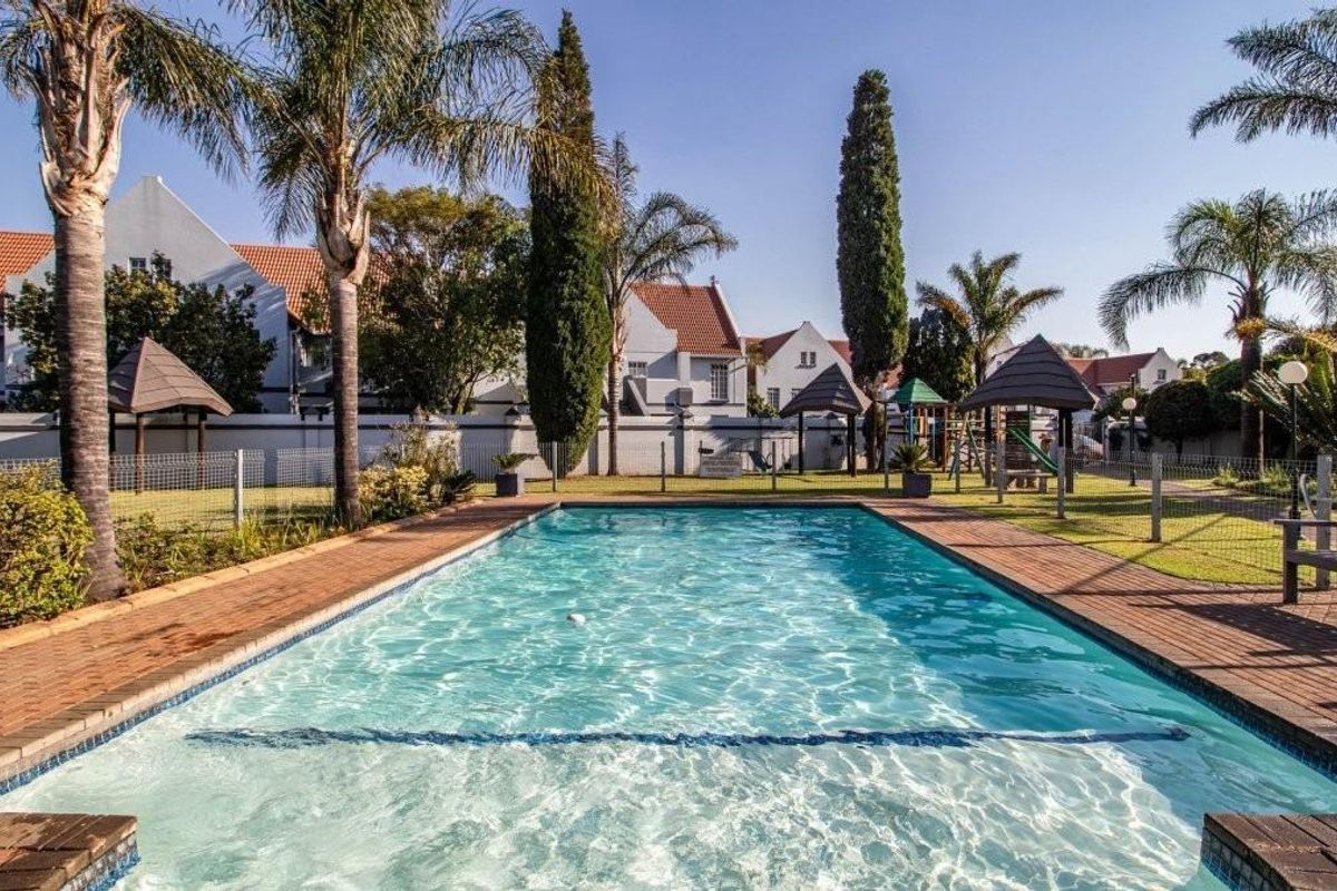 A well-maintained communal swimming pool area featuring brick paving, landscaped gardens, and mature palm trees within a secure townhouse complex.