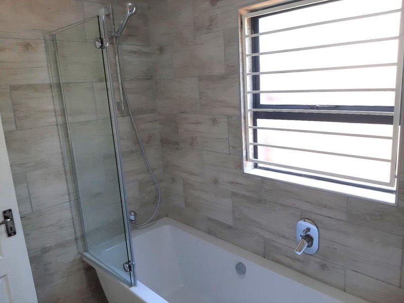 A clean, modern bathroom featuring a bathtub with a glass shower screen, neutral wood-look wall tiles, and a window with security bars.