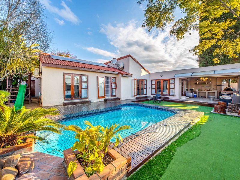 A well-maintained outdoor entertainment area featuring a swimming pool with wooden decking, an artificial lawn, and a covered patio with a built-in kitchen.