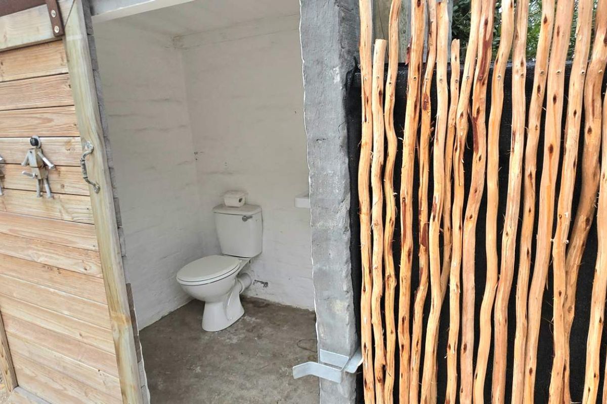 An outdoor rustic toilet facility featuring a simple porcelain toilet bowl, timber door, and natural wood pole privacy screening.