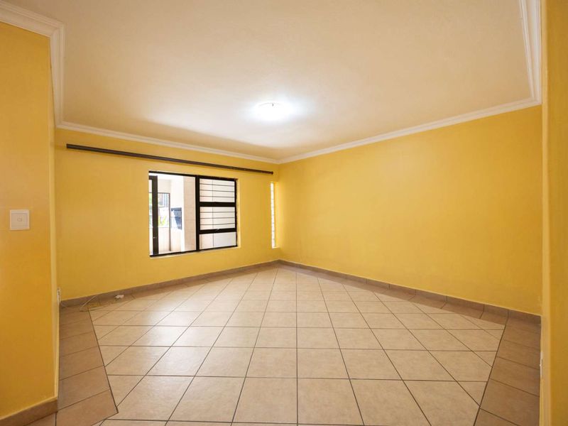 A spacious, empty living area featuring neutral tiled flooring, yellow painted walls, and aluminium-framed windows with security bars.