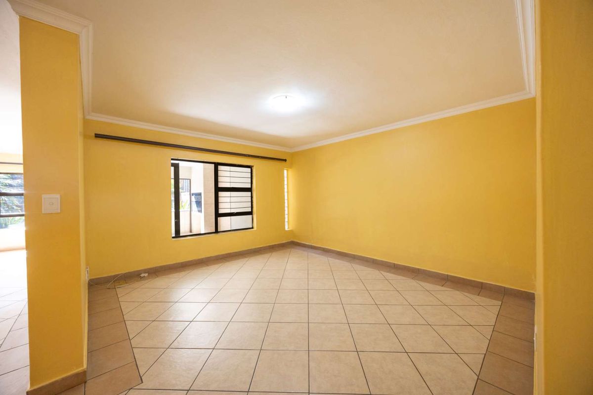 A spacious, empty living area featuring neutral tiled flooring, yellow painted walls, and aluminium-framed windows with security bars.