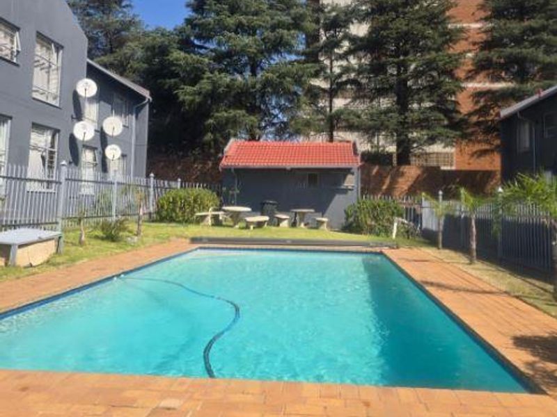 Outdoor communal pool area featuring a rectangular swimming pool, brick paved surround, and metal perimeter fencing within an apartment complex setting.