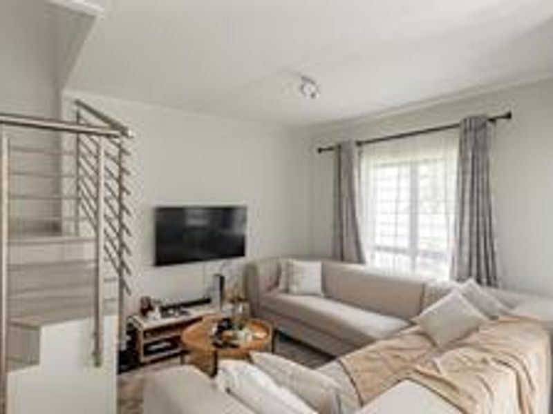 A bright living room featuring a staircase with a modern stainless steel balustrade, tiled flooring, and large windows with curtain rails.