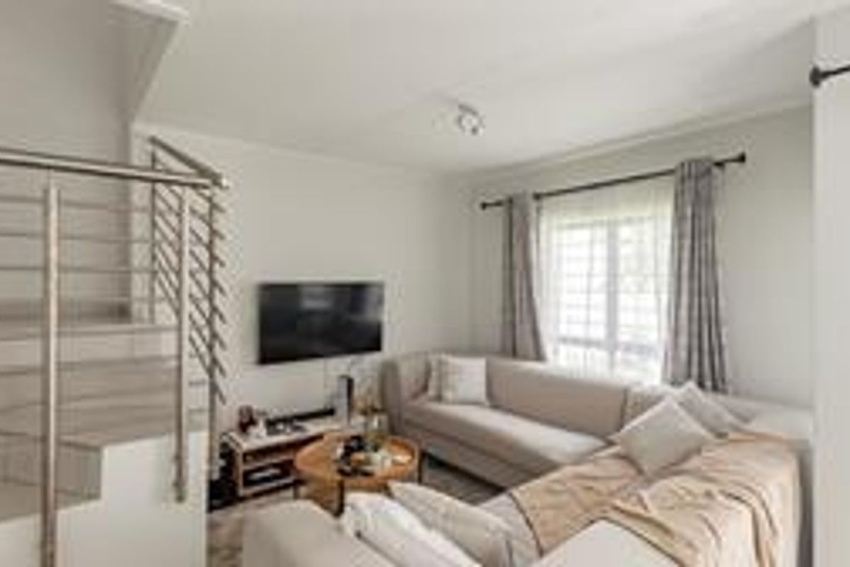 A bright living room featuring a staircase with a modern stainless steel balustrade, tiled flooring, and large windows with curtain rails.