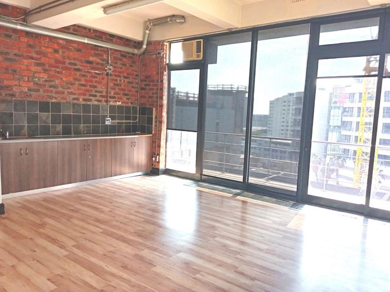 Open-plan office space featuring exposed brick walls, laminate flooring, a small kitchenette area, and large windows providing ample natural light and balcony access.