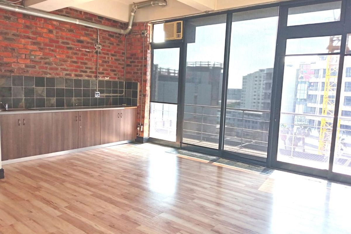 Open-plan office space featuring exposed brick walls, laminate flooring, a small kitchenette area, and large windows providing ample natural light and balcony access.