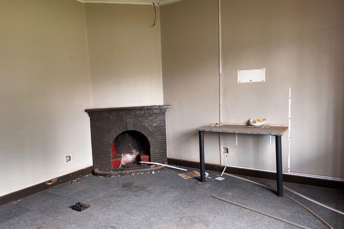 An empty living room featuring a traditional brick fireplace, crown molding, and worn carpeting, requiring cosmetic updates and electrical repairs.