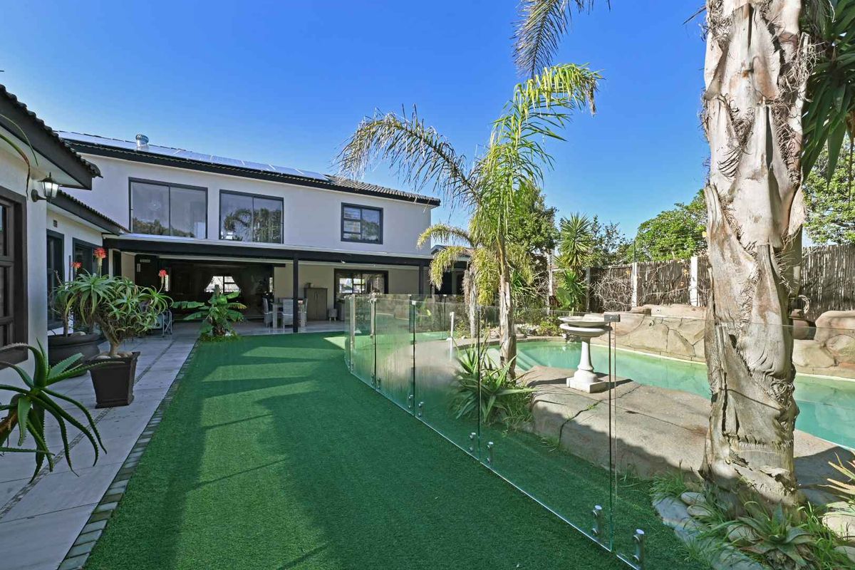 Rear exterior view featuring a swimming pool with glass fencing, artificial turf lawn, paved patio area, and solar panels on the roof.