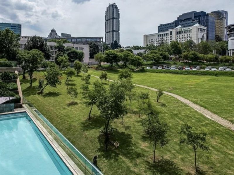 An elevated view of a swimming pool with glass balustrades and timber decking, overlooking a landscaped park and urban skyline.