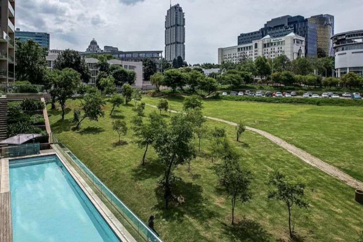 An elevated view of a swimming pool with glass balustrades and timber decking, overlooking a landscaped park and urban skyline.