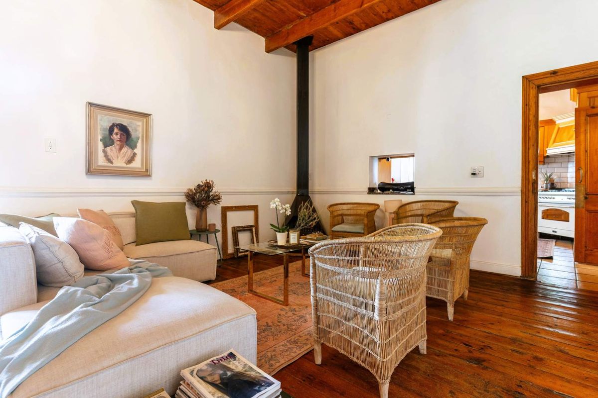 A cozy living room featuring exposed timber ceiling beams, a central fireplace, and original hardwood flooring, with a view into the adjacent kitchen.