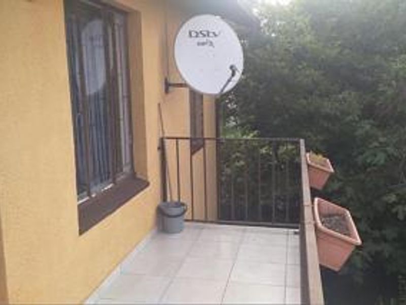 A compact balcony featuring tiled flooring, a metal balustrade, and a wall-mounted satellite dish, overlooking surrounding greenery.