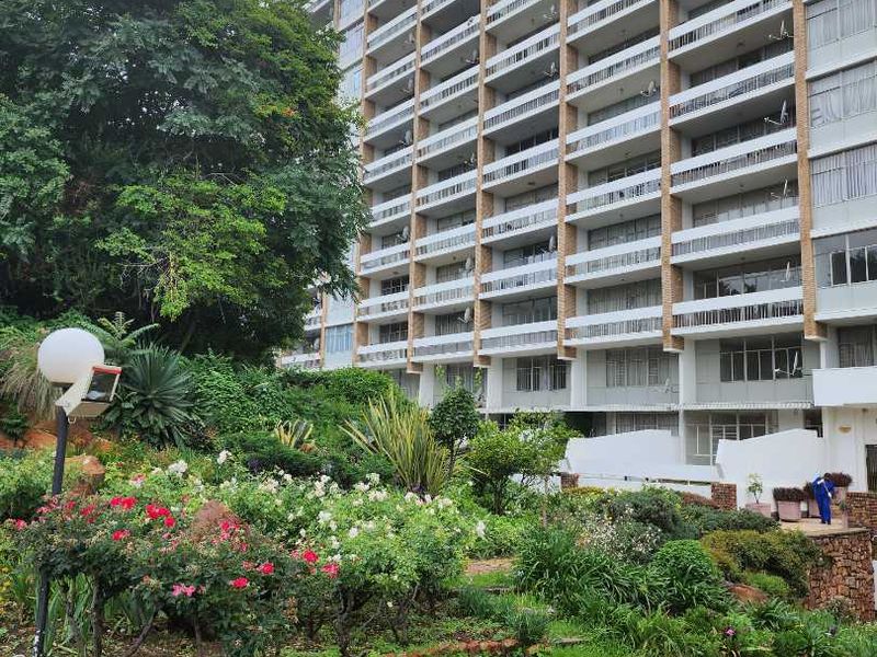 Exterior view of a multi-story apartment building featuring private balconies and a landscaped garden area with a paved driveway.
