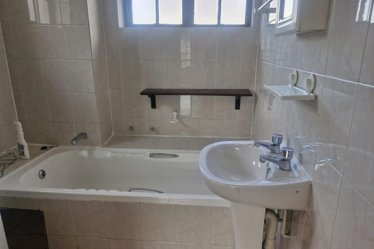A functional bathroom featuring a standard bathtub, pedestal basin, and full-height wall tiling with a window for natural light.