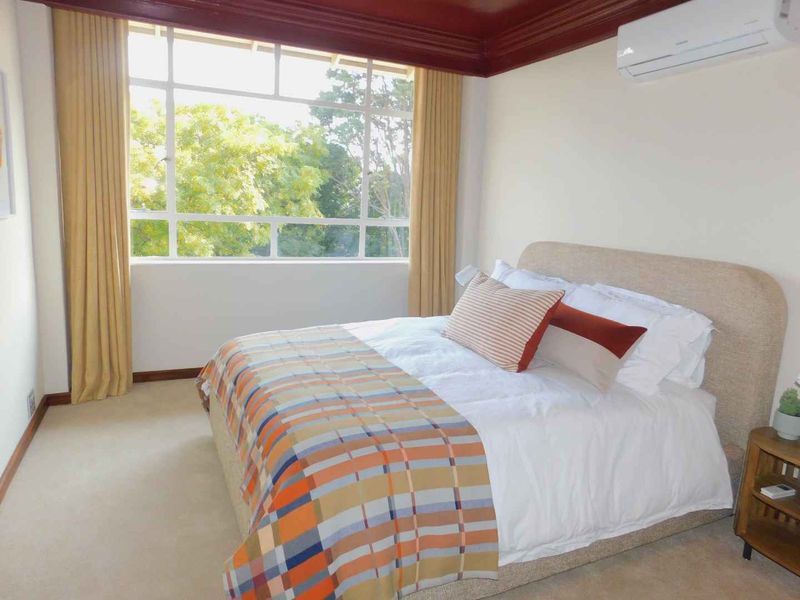 A bright and comfortable bedroom featuring carpeted flooring, a split-system air conditioner, and a large window providing natural light and pleasant outdoor views.