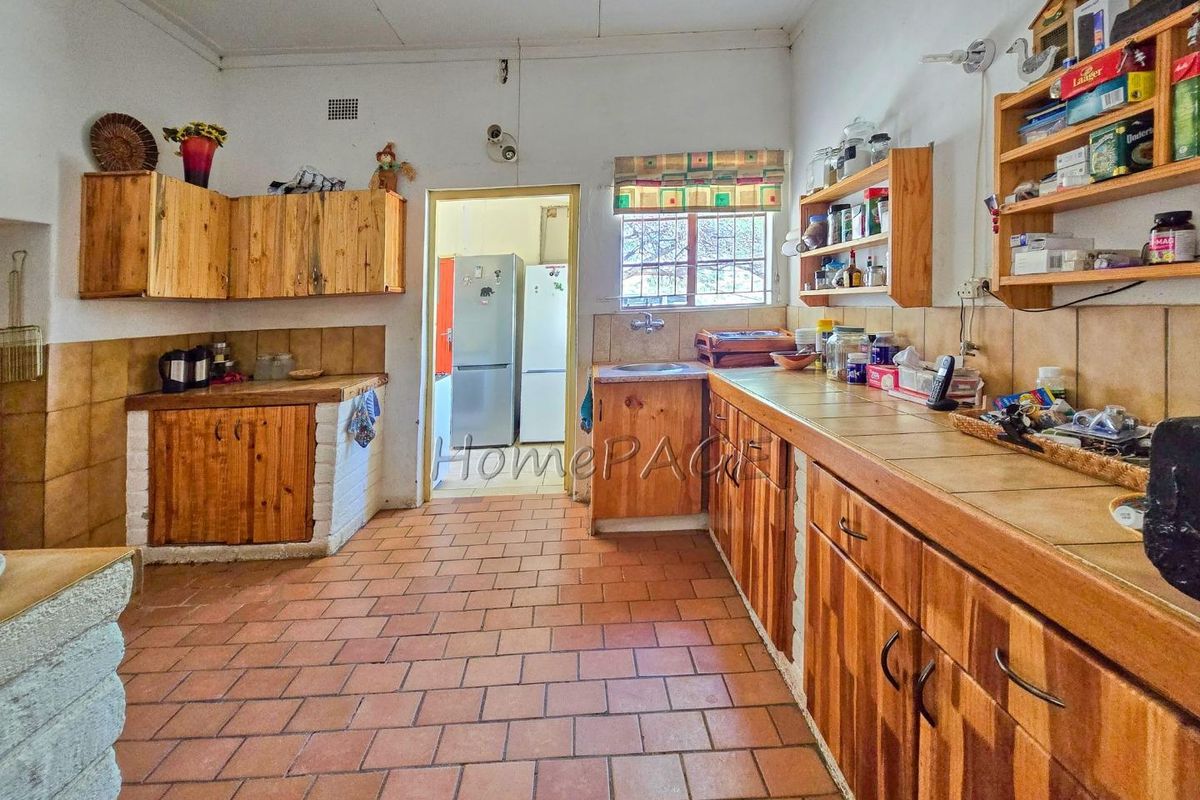 A rustic kitchen featuring wooden cabinetry, tiled countertops, and terracotta floor tiles, offering a functional layout with direct access to an adjacent utility area.