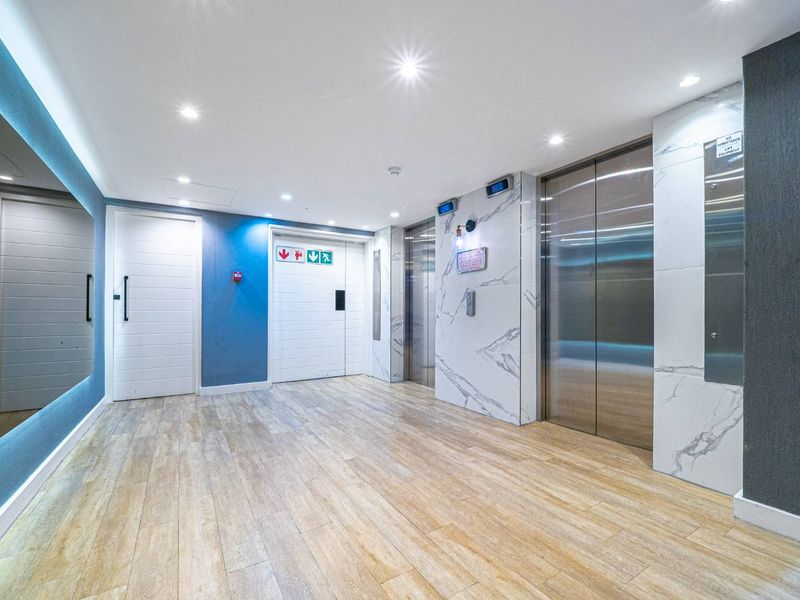 Modern apartment building lobby or hallway featuring marble-effect wall cladding, stainless steel elevator doors, and wood-look flooring with recessed lighting.