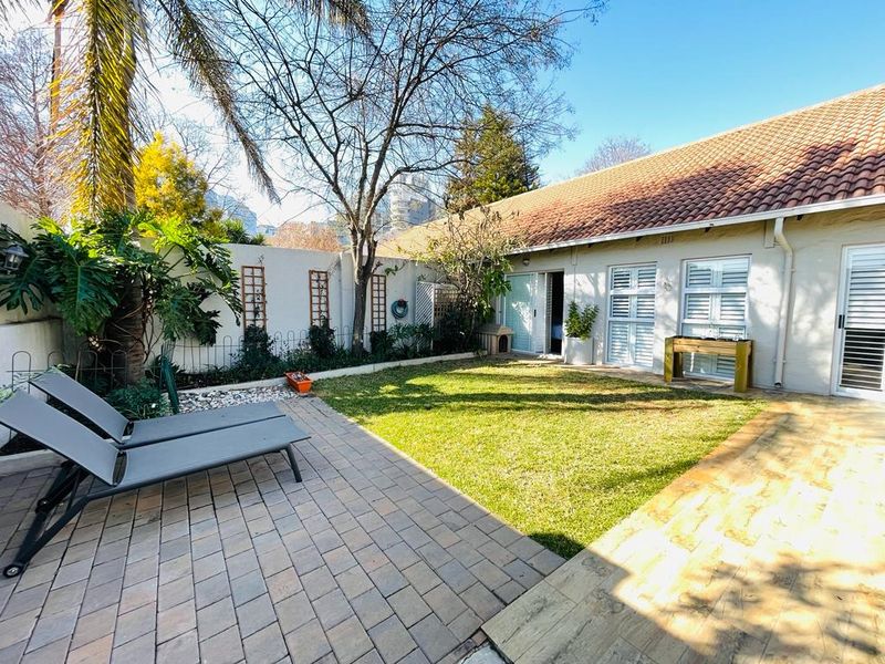 A private rear garden featuring a paved patio area, well-maintained lawn, and direct access to the townhouse via sliding doors and shuttered windows.