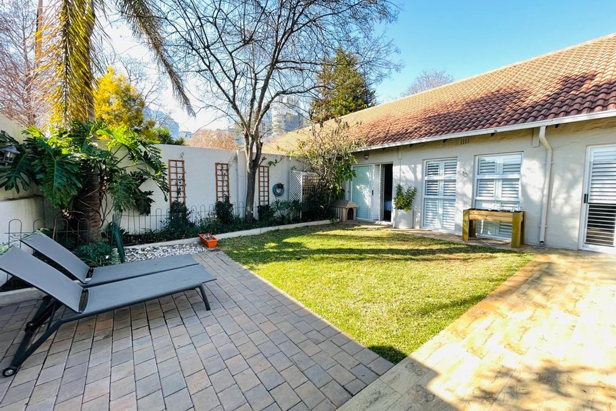 A private rear garden featuring a paved patio area, well-maintained lawn, and direct access to the townhouse via sliding doors and shuttered windows.